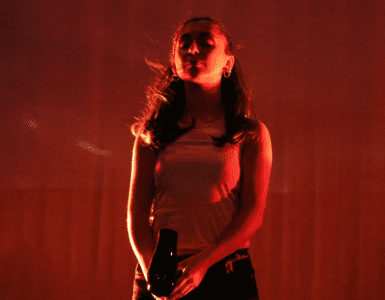 Dark red stage lighting. A young woman stands straight, eyes closed, blowing a hairdryer into her face.