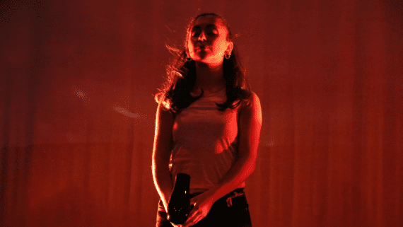 Dark red stage lighting. A young woman stands straight, eyes closed, blowing a hairdryer into her face.