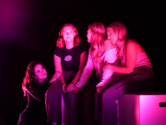 A photo of four women bathed in pink light with their arms around each other, smiling.