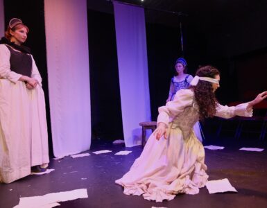 Lady Jane Grey sits blindfolded in white, reaching out ahead of her. Two 16th century dressed young women look on.