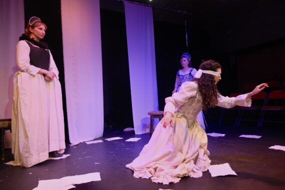 Lady Jane Grey sits blindfolded in white, reaching out ahead of her. Two 16th century dressed young women look on.