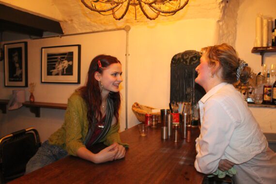 Two women In a pub, lean over the bar chatting.